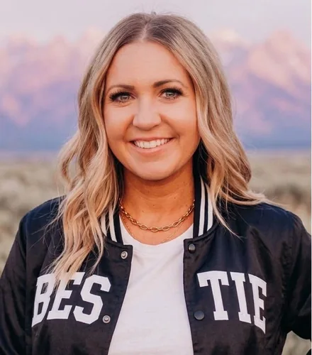 Dani, founder of the Bestie Squad, smiling in a black Bestie bomber jacket in front of the mountains.