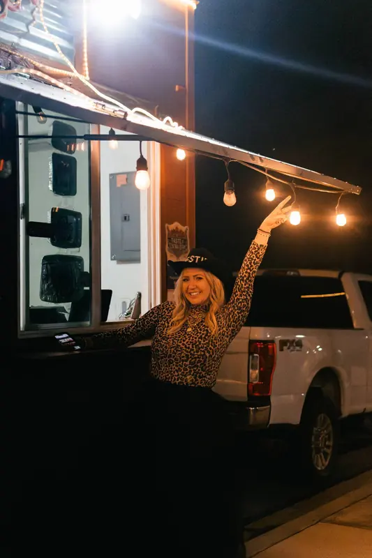 Dani, founder of the Bestie Squad, at night by a lit food-truck window under string lights, in a black cowboy hat with one arm raised.